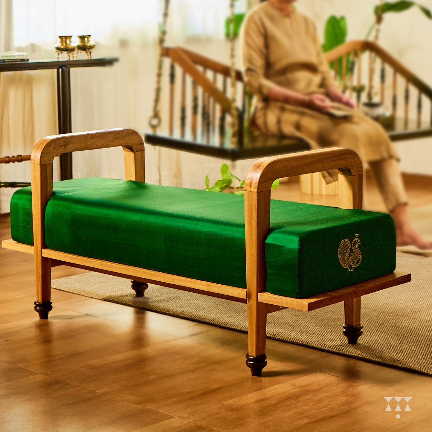 Green cushioned bench with wooden frame on a wooden floor, person sitting on a swing in the background.